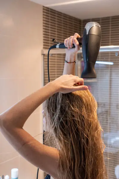 Blonde woman with wet hair drying her hair with dryer in modern bathroom in front mirror
