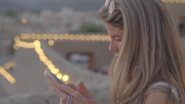 Sarışın bir kadın yolcunun profilinin yatay videosu. Akşamları terasta Müslüman bir şehrin manzarası eşliğinde cep telefonunu kaydırıyor.