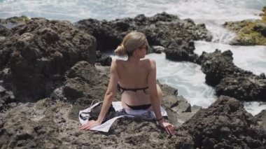 Sarışın bir kadının, denizin kenarında kayaların üzerinde güneşlenirken gerçek zamanlı görüntüsü.