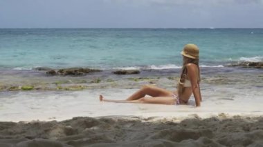 Bikini ve hasır şapkalı güzel bir kadının denizin kenarında güneşlenirken çekilmiş gerçek zamanlı yatay videosu.