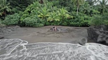 Kafkasyalı bir çiftin ve köpeğinin kumlu, sade bir plajda oturduğu, siyah kum ve palmiye ağaçlarının olduğu bir hava görüntüsü. Nuqui bölgesi, Kolombiya