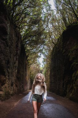 Bir kadın ormanda bir patikada yürüyor. Beyaz bir gömlek ve şort giyiyor.