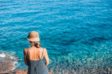 Güneş şapkası takan bir kadın kayalık zeminde duruyor ve berrak mavi okyanusa bakıyor. Parlak güneş ışığı, yansıma için mükemmel, sakin bir atmosfer yaratır..
