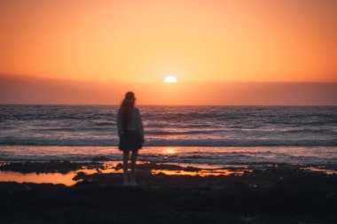 Defocused person stands alone on the beach as the sun sets, casting warm hues across the sky and water. The tranquil scene captures a moment of solitude and reflection. clipart