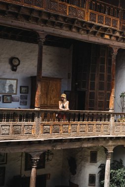 Bir kadın, karmaşık ahşap oymalarla bezenmiş bir balkonda huzur içinde oturur. Eski moda dekorlar ve tarihi bir binanın sıcak, davetkar atmosferiyle çevrili..