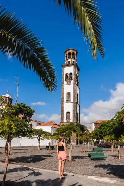 Büyüleyici bir kasaba meydanında duran bir insan, tarihi bir kuleye hayran. Parlak gökyüzü ve yemyeşil palmiye ağaçları keşif için rahatlatıcı bir atmosfer yaratır. Kanarya Adaları, Tenerife