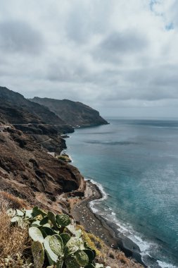Panoramik bir manzara kıyıya vuran yumuşak dalgalarla kayalık kıyı şeridini yakalar, yemyeşil ve gökyüzündeki dramatik bulutlar tarafından çerçevelenir..