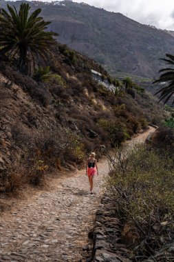 Pembe etekli bir kadın kayalık bir yolda yürüyor, canlı bitki örtüsü ve dik tepelerle çevrili, bulutlu bir gökyüzünün altında, sakin bir dağda..