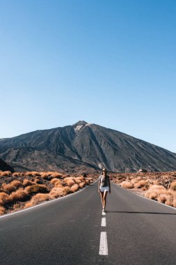 Bir kadın Teide, Tenerife, Kanarya Adaları 'nın önünde yolda yürüyor. Gökyüzü açık ve güneş parlıyor.