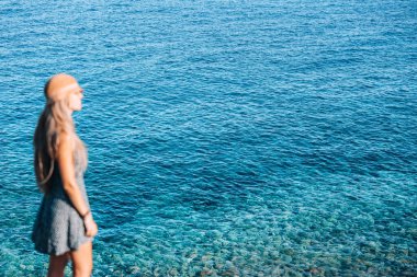 Berrak turkuaz suların yanında duran, dağılmış bir kadın, açık güneşli bir günde, kayalık bir sahil şeridiyle çevrili, sakin okyanus manzarasının tadını çıkarıyor..