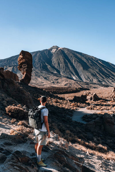 Hiker man stands on rugged terrain, admiring the towering mountain backdrop in Teide National Park, surrounded by unique rock formations and clear blue skies.