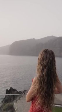 Tenerife 'deki Los Gigantes tepelerinin ardında güneşin batışı gibi huzurlu kıyı manzarasına hayran bir şekilde kayalık bir arazide duran kadının arka görüntüsü. Ortalık sakin.