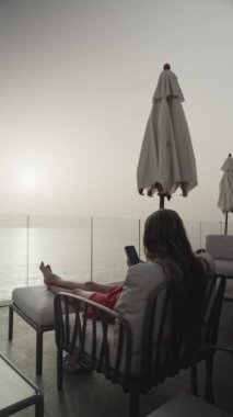 Bir kadının deniz kenarındaki bir tatil köyünde sakin bir anın keyfini çıkarırken çekilmiş dikey videosu. Sandalyeye uzanmış ve güneş suyun üzerinde batarken akıllı telefonuna bakıyor..