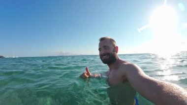 Yavaş çekim Vlog adam güneşli bir sahilde açık okyanusta mutlu bir şekilde yüzüyor, açık havanın tadını çıkarıyor..