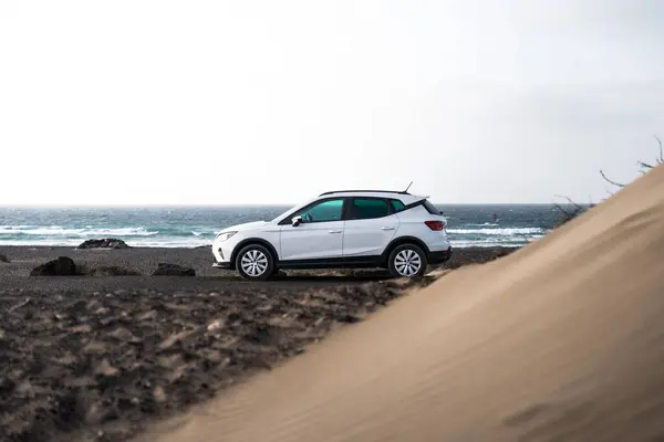 Famara kıyısı boyunca kumlu kum tepelerinin yanına park etmiş beyaz bir araba. Lanzarote 'de bulutlu bir günde arka planda dalgalar çarpıyor.