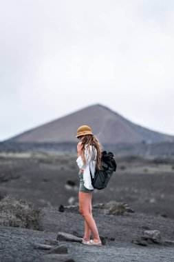 Sarışın kadın nükteli şapkası, çorak bir volkanik alanda durup sırt çantası takmış, uzak bir dağa bakıyor..
