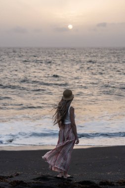 Gün batımında bir kadın sahilde yürüyor, dalgalar yavaşça ayaklarına vuruyor..