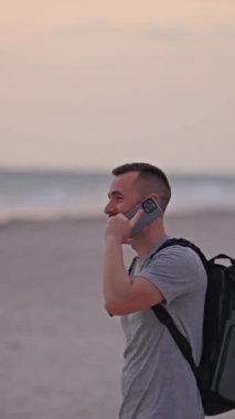 Bir adam kumsalda sırt çantasıyla duruyor, gün batımının tadını çıkarırken bir telefon görüşmesi yapıyor..