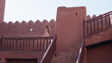 Umman 'daki eşsiz Arap mimarisi ve kültürünün tadını çıkaran bir kadın parlak güneşin altındaki tarihi kerpiç yapılarından geçiyor.