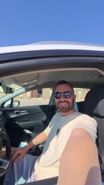 Güneşli bir günün tadını çıkaran bir adam, arabasında oturup, güneş gözlüğü takarken büyük bir gülümseme sergiliyor. Vlog
