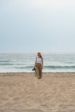 Kumlu bir sahilde elinde fotoğraf makinesi ve haritasıyla yürüyen bir kadın fotoğrafçı.