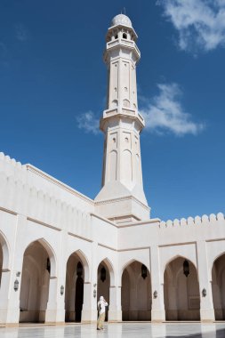 Müslüman bir kadın, mavi gökyüzünün altında uzun bir minareyle beyaz, modern bir caminin avlusunda dua ediyor.