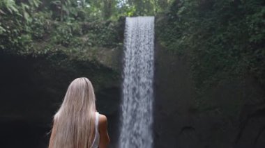Turistin göz kamaştırıcı şelalenin yemyeşil ormandaki yosunlu kayalıklardan düşüşünü dikey olarak izlemesi.