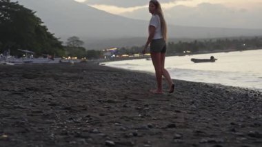 Genç bir kadın alacakaranlıkta parlayan siyah kumsalda yalınayak yürüyor. Arka planda agung Dağı var.