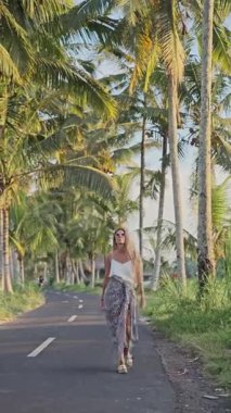 Sarışın kadın Endonezya, Bali 'de palmiye ağaçlarıyla dolu bir yolda yürüyor.