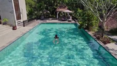 Beyaz bikinili bir kadın tropik bitki örtüsüyle çevrili güzel bir havuzda yüzüyor.