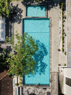 Tropik bir tatil beldesinde turkuaz suyu bulunan dikdörtgen yüzme havuzunun havuz kenarında havuz kenarında oturan bir turist görülüyor.