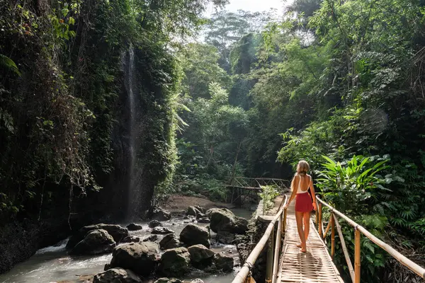 Genç kadın turist bambu köprüsünde yürüyor ve tropikal orman ormanlarında güzel şelalenin tadını çıkarıyor.