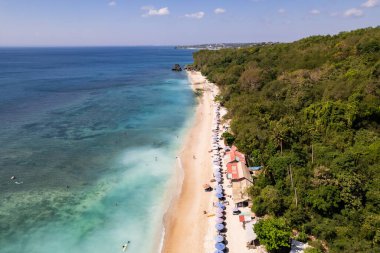 Bali, Endonezya 'da güzel bir tropik sahil manzarası, turkuaz su, plaj şemsiyeleri ve yemyeşil bitki örtüsünü gösteriyor.
