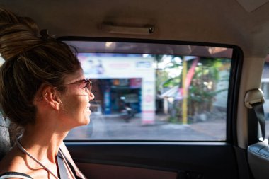 Mutlu bir bayan turist arabasının camından dışarı bakıyor, Balis 'in canlı kültürünün tadını çıkarıyor.
