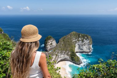 Hasır şapkalı sarışın turist Nusa Penida, Bali 'deki uçurum manzaralı Kelingking plajına hayran.