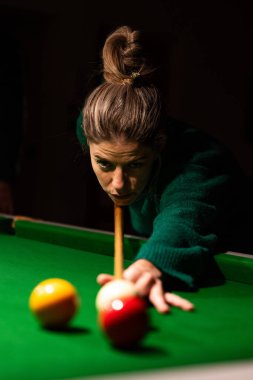 Yeşil bir bilardo masasına bilardo topu doğrultan odaklanmış kadın oyuncu, loş bir ayarda konsantrasyon ve beceri gösteriyor.