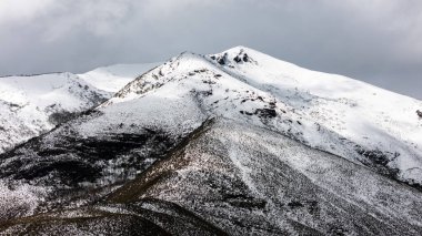 Karla kaplı görkemli dağ zirveleri bulutlu bir gökyüzünün altındaki ufka hükmediyor ve nefes kesici bir kış manzarası yaratıyor.