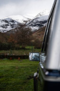 Park halindeki off-road aracı karlı dağ manzarasını yansıtıyor. Yağmurla kaplı yan aynada, macera ve keşifler sergiliyor.