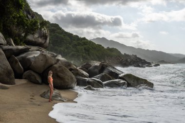 Sarışın turist bulutlu bir günde Kolombiya, Tayrona Ulusal Parkı plajında dalgaların ve kayaların keyfini çıkarıyor.