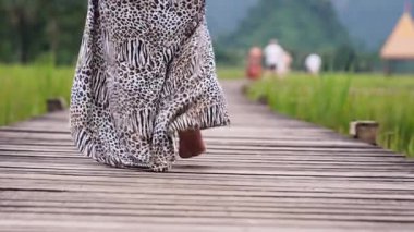 Low-angle shot of a woman in a long dress walking barefoot on a wooden path through a green field