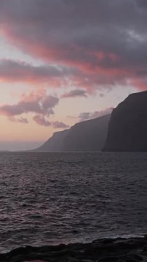 Calming ocean waves are crashing on a rocky shore with dramatic cliffs in Los Gigantes, Tenerifeunder a colorful twilight sky