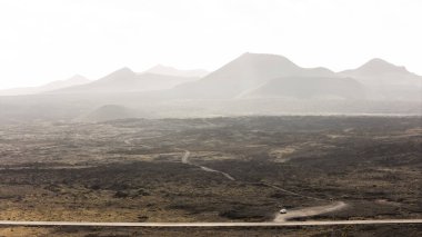 A lone vehicle travels a dusty road through a vast, barren landscape of distant mountains under a cloudy sky in Lanzarote, Canary Islands