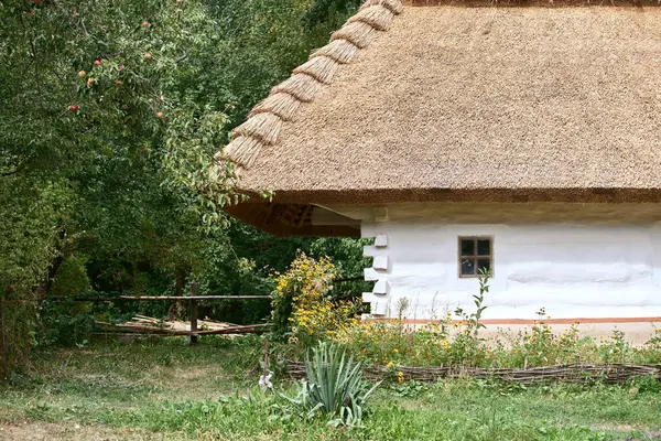 stock image Part of an old clay house with a wooden brown window and a thatched roof. Traditional Ukrainian village house with garden and wooden fence