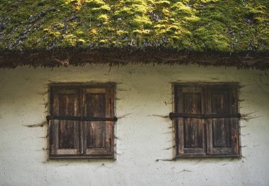 Ahşap pencereli, tahta çatılı, yosun ve liken kaplı terk edilmiş eski bir evin parçası. Kil bir evin beyaz duvarında kapalı kepenkleri olan iki kahverengi pencere.