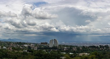 Bu panoramik manzara doğa ve şehir yaşamı arasındaki sakin zıtlığı yakalar. Yükselen kümülüs bulutları uçsuz bucaksız mavi gökyüzü boyunca uzanır, yuvarlanan yeşil tepeler ve uzak şehir silueti üzerine dinamik gölgeler bırakır. Ön planda, yemyeşil sebzeler