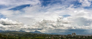 Bu panoramik manzara doğa ve şehir yaşamı arasındaki sakin zıtlığı yakalar. Yükselen kümülüs bulutları uçsuz bucaksız mavi gökyüzü boyunca uzanır, yuvarlanan yeşil tepeler ve uzak şehir silueti üzerine dinamik gölgeler bırakır. Ön planda, yemyeşil sebzeler