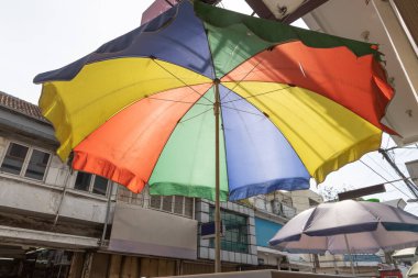 Kalabalık bir sokak pazarında büyük renkli bir şemsiye gölge sağlar. Aşağıdan çekilen, kırmızı, sarı, yeşil ve mavi paneller güneş ışığı altında parlıyor..