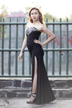 Sofistike siyah straplez gece elbisesi içinde dengeli bir kadın, korse üzerinde göz kamaştırıcı detaylar ve şık kalçalı bir kesik. Elini kalçasında gezdirip kendine güvenen bir duruş sergiliyor. Elbisenin zarif siluetini gösteriyor. 