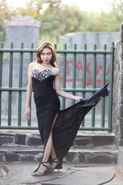 Sofistike siyah straplez gece elbisesi içinde dengeli bir kadın, korse üzerinde göz kamaştırıcı detaylar ve şık kalçalı bir kesik. Elini kalçasında gezdirip kendine güvenen bir duruş sergiliyor. Elbisenin zarif siluetini gösteriyor. 