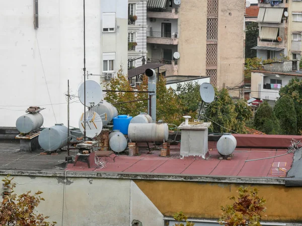 stock image Cisterns, water tanks and satellite dishes on a planted roof of a building in Tirana. In the background, the city surrounded by mountains.